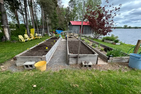 Raised garden beds with soil, surrounded by grass and trees, with a red-roofed building and a body of water in the background.